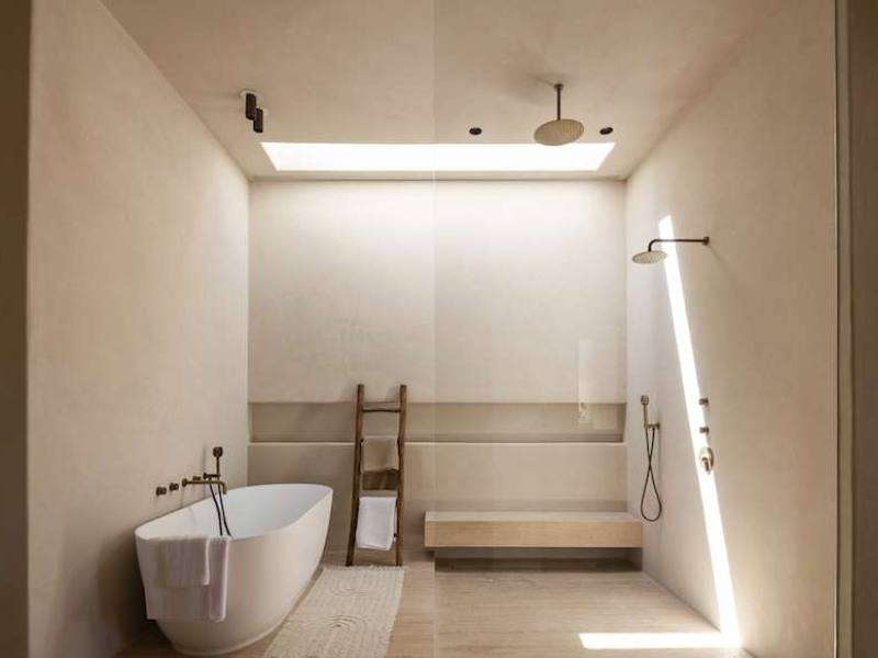Open-concept bathroom finished in Meoded Concretta plaster across walls and ceiling, featuring a seamless design with a soaking tub beside a plastered walk-in shower, all softly illuminated by natural light from a skylight above.