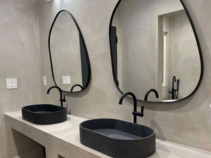 Ultra-modern bathroom finished in Meoded Concretta plaster in a soft grey tone, providing a subtle, refined backdrop to bold black sinks, matte black hardware, and sculptural, organic-shaped designer mirrors.